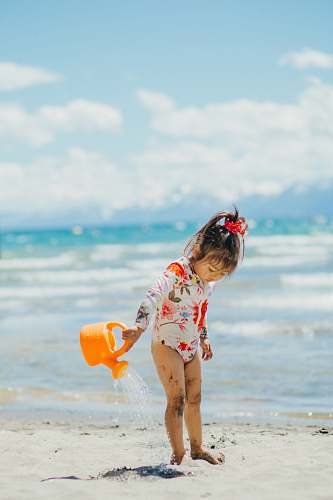 kids swimming girl in white and pink floral shirt holding orange plastic bucket on beach during daytime girl alone